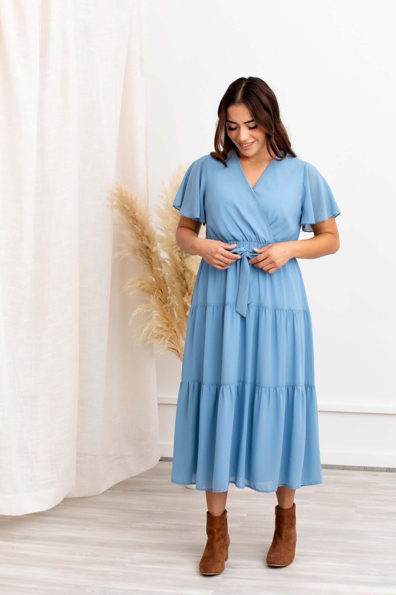 A woman wearing a blue midi dress with short, fluttery sleeves and a V-shaped neckline. The dress features a tiered skirt and a waist tie.