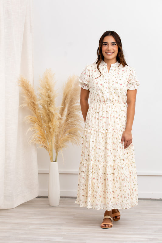 A woman standing in front of a plant decoration, wearing a beige floral cotton maxi dress with short puffed sleeves, a round neckline, and a tiered skirt with ruffles.