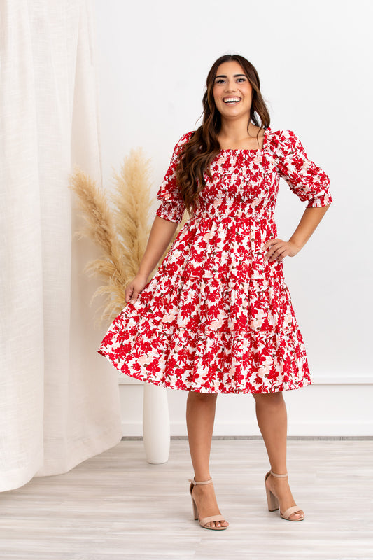 A woman standing in a flowery indoor setting wearing a red floral mini dress with a square neckline and puff sleeves.