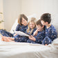 Four children in matching pajamas sitting on a bed reading a book, with a decorated Christmas tree in the background.