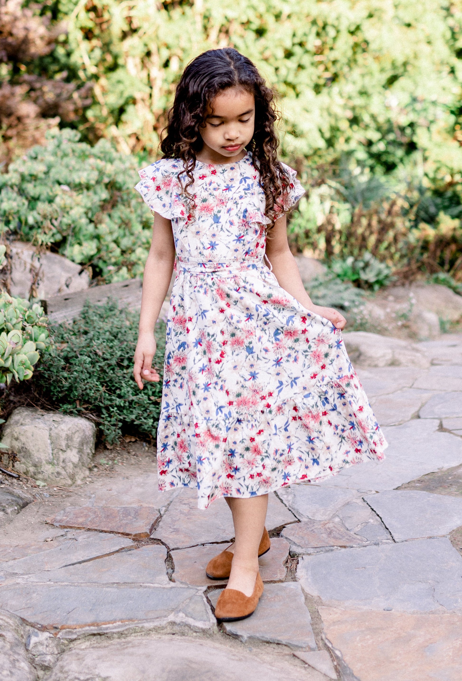 Young girl in a floral dress standing on a stone path with greenery in the background