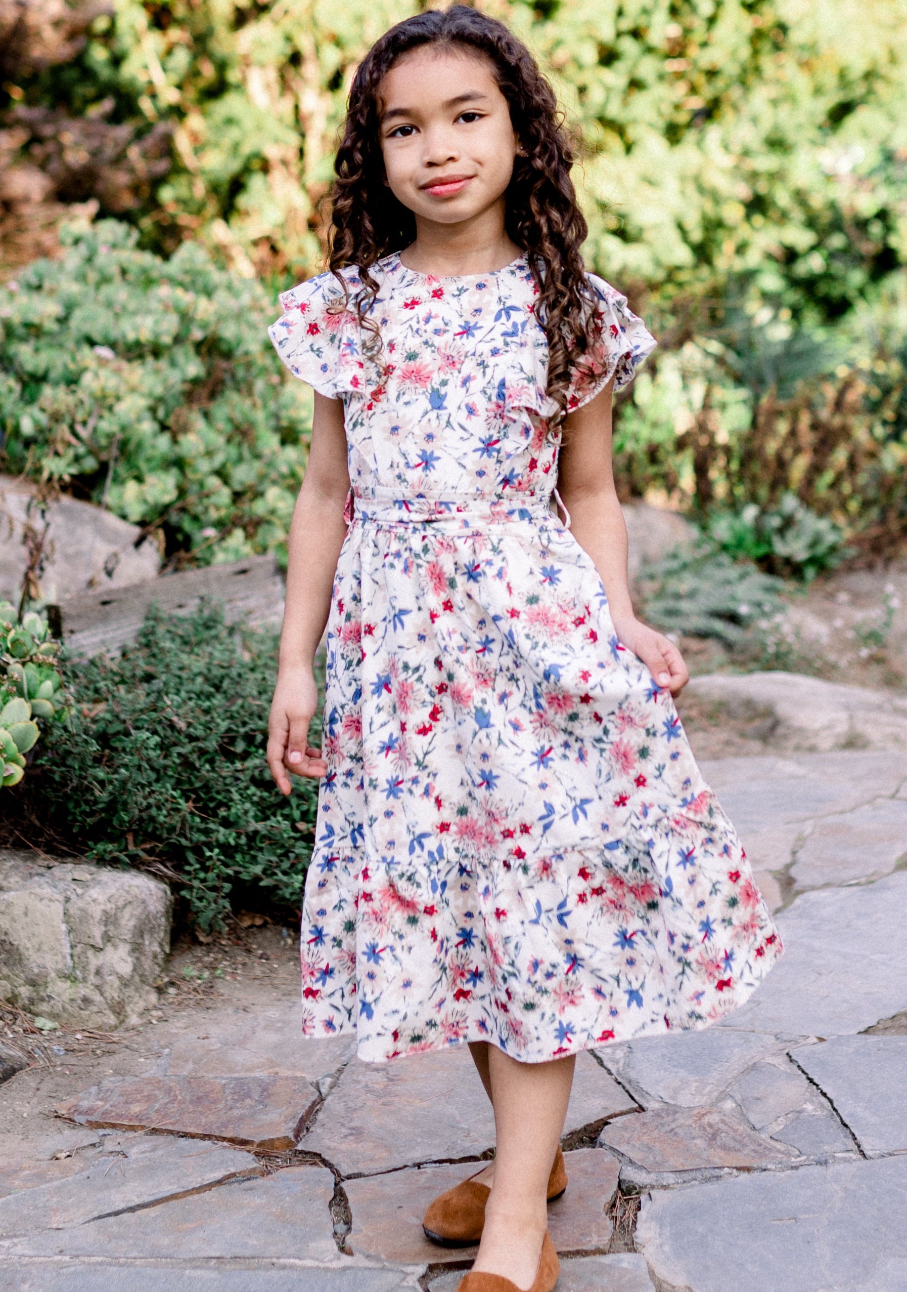 Young girl in a floral dress standing on a stone path in a garden.