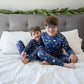 Two children in matching pajamas sitting on a bed with a festive headboard.