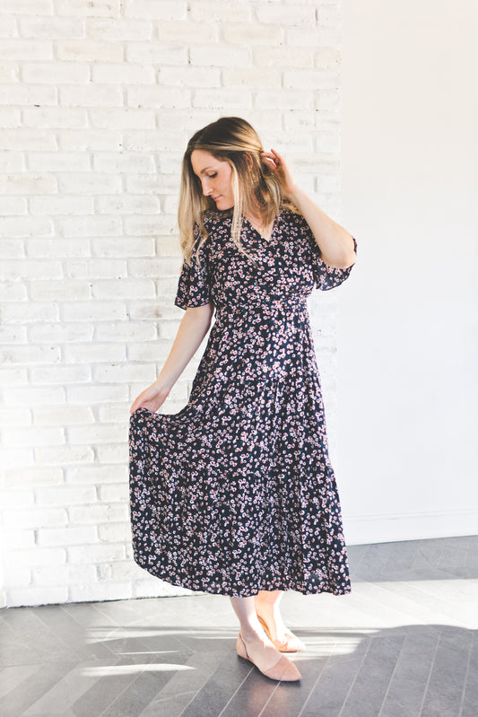 A woman wearing a navy floral print midi dress with flutter sleeves and a V-neckline, standing against a white brick wall.