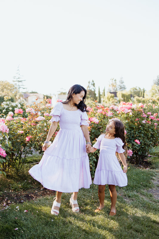 A woman and a young girl wearing matching lilac dresses with short sleeves and knee-length, standing in a garden.