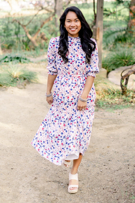 A woman standing in a natural setting wearing a white floral midi dress with three-quarter sleeves and a cinched waist.