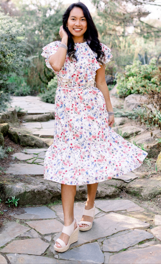 A woman standing in a garden setting, wearing a white floral midi dress with ruffle sleeves and a high neckline.