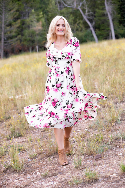 A woman standing in a field, wearing a pink floral midi dress with half sleeves and a tiered skirt.