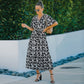 A woman wearing a floral midi dress with a V-neckline, short sleeves, and a belt at the waist. The dress is predominantly blue with a pattern of smaller white flowers.